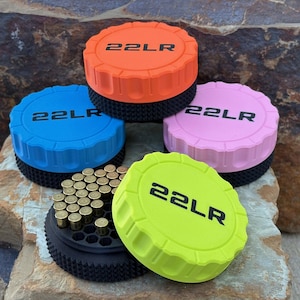 May include: Four circular ammunition storage containers with black textured bases and lids in orange, blue, pink, and neon yellow. Each lid features the text 22LR in black. One container is open, revealing a black honeycomb tray holding brass cartridges.