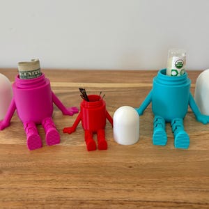 以下が含まれることがあります： 腕と脚のあるフィギュアの形をした、3つのカラフルで気まぐれな容器。1つはマゼンタで中に紙幣が入っており、もう1つは赤で小さなアイテムが入っており、3つ目は小さなチューブが入ったティールです。それぞれに白い蓋が付いています。