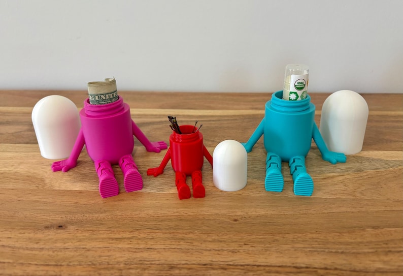 以下が含まれることがあります： 腕と脚のあるフィギュアの形をした、3つのカラフルで気まぐれな容器。1つはマゼンタで中に紙幣が入っており、もう1つは赤で小さなアイテムが入っており、3つ目は小さなチューブが入ったティールです。それぞれに白い蓋が付いています。