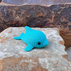May include: A turquoise knitted dolphin toy with black eyes. The dolphin is lying on a light brown rock, with a large, textured rock in the background. The toy has a textured surface and is made of a soft material.