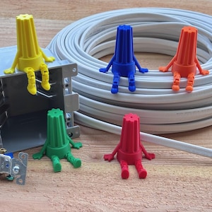 May include: A collection of colorful wire connectors, each with arms and legs, arranged around a roll of white electrical wire. The connectors are yellow, blue, orange, green, and red. An electrical box and a light switch are also visible.