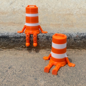 May include: Two orange and white traffic cone figurines with arms and legs are sitting on a concrete surface. The cones have a cylindrical shape with white stripes and a handle on top. The figurines are in a relaxed pose.