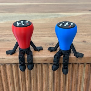 May include: Two novelty shift knobs, one red and one blue, each with black legs and arms. The knobs display a gear shift pattern on a black surface. The items are sitting on a wooden surface.
