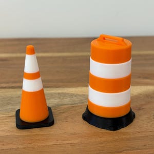 May include: A miniature orange and white traffic cone with a square black base next to a cylindrical orange and white traffic barrel featuring a handle on top and a circular black base. Both items are constructed with alternating horizontal stripes.