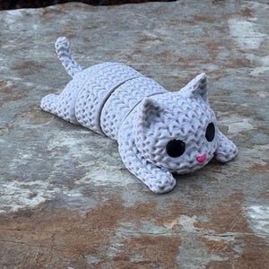 May include: A light gray cat figurine with a textured surface, black eyes, and a pink nose. The cat is lying down with its tail curved. The figurine is made of a segmented material and is resting on a stone surface.