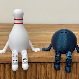 May include: A white bowling pin with red stripes and a blue bowling ball with arms and legs sit side-by-side on a wooden surface. Both have articulated limbs, giving them a playful appearance. The objects are likely 3D-printed.