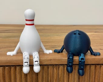 May include: A white bowling pin with red stripes and a blue bowling ball with arms and legs sit side-by-side on a wooden surface. Both have articulated limbs, giving them a playful appearance. The objects are likely 3D-printed.