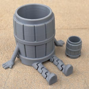 May include: A gray 3D printed figurine of a barrel with legs and arms. The barrel is sitting on a light brown surface.