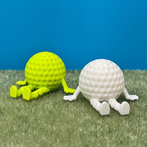 May include: Two whimsical golf ball figurines, one neon green and one white, each with arms and legs, resting on artificial turf against a blue backdrop. The golf balls have a dimpled surface.