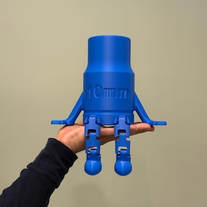May include: A bright blue, 3D-printed object with the text "10mm" on its side. The object has a cylindrical body, two arms, and two legs. The legs are articulated and end in rounded feet. The object is held in a person's hand.