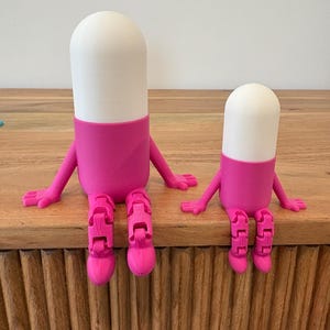 以下が含まれることがあります： 2つの気まぐれなピル型の置物。それぞれ白い上部と鮮やかなピンク色の中央部、腕、関節のある脚があります。まるで座っているかのようにポーズをとっており、デザインに遊び心を加えています。置物は木製の表面に展示されています。