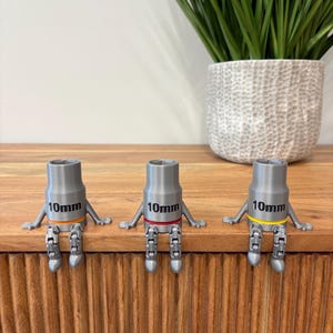 May include: Three gray 10mm socket figures with articulated legs and arms, sitting on a wooden surface. Each figure has a different colored band: orange, red, and yellow. A decorative plant in a white pot is in the background.