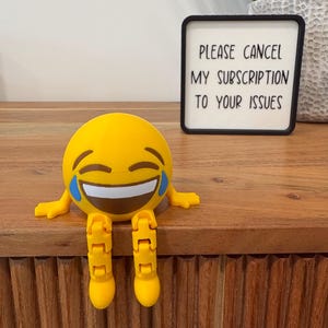 May include: A yellow 3D printed emoji figure with a laughing expression, blue tears, and articulated dangling legs. Next to it is a black square sign with white text that reads: PLEASE CANCEL MY SUBSCRIPTION TO YOUR ISSUES in black lettering.
