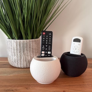 May include: A white and a black textured spherical remote control holder, each holding a remote. A green plant in a textured white pot is in the background. The remote control holders are on a wooden surface.
