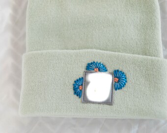Puede incluir: Un gorro verde claro con un borde doblado. El borde presenta un parche cuadrado gris con tres flores azules bordadas con centros naranjas. El gorro está hecho de un material suave y tejido.