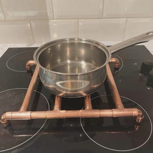 May include: A stainless steel saucepan with a long handle resting on a square trivet constructed from copper pipes and fittings. The trivet features four corner legs and a grid-like frame design, providing a raised base for the metal cookware.