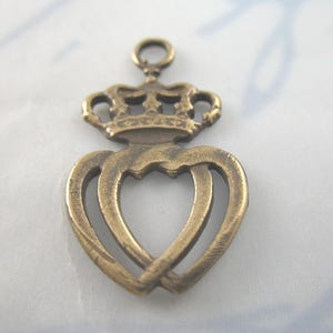 May include: A bronze-colored charm featuring a crown above two intertwined hearts. The charm has a loop at the top for attaching to a necklace or bracelet. The design has a vintage, antique appearance.