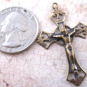 May include: An antique bronze-toned crucifix pendant with a detailed depiction of Jesus Christ. The cross is ornate with decorative flourishes. A U.S. quarter coin is next to the cross for size comparison. The coin is dated 1980.