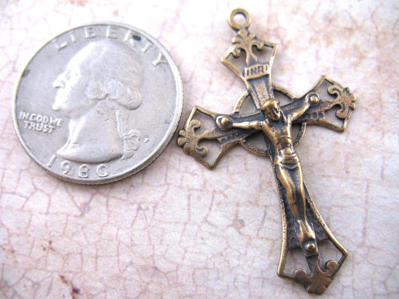 May include: An antique bronze-toned crucifix pendant with a detailed depiction of Jesus Christ. The cross is ornate with decorative flourishes. A U.S. quarter coin is next to the cross for size comparison. The coin is dated 1980.