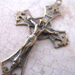May include: A detailed bronze crucifix pendant. The cross features an ornate design with a figure of Jesus Christ. The pendant has a loop at the top for hanging and is likely made of metal. The overall style is vintage or antique.