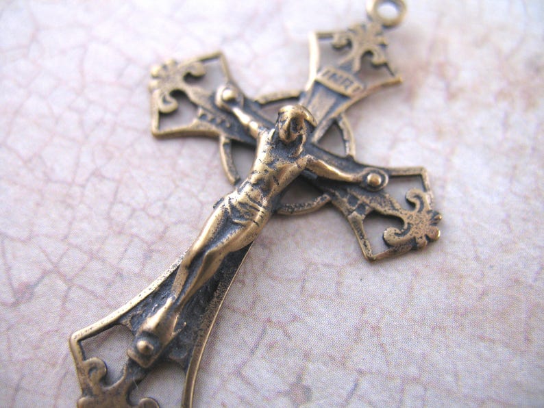 May include: A detailed bronze crucifix pendant. The cross features an ornate design with a figure of Jesus Christ. The pendant has a loop at the top for hanging and is likely made of metal. The overall style is vintage or antique.