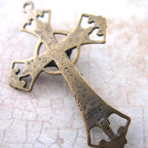 May include: A detailed, antique-style cross pendant in a bronze finish. The cross features ornate cut-out designs on the arms and a circular element at the center. The metal has a slightly aged, textured appearance, suggesting a vintage aesthetic.