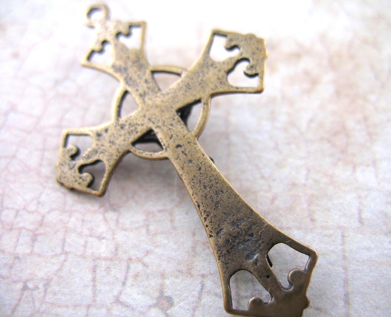 May include: A detailed, antique-style cross pendant in a bronze finish. The cross features ornate cut-out designs on the arms and a circular element at the center. The metal has a slightly aged, textured appearance, suggesting a vintage aesthetic.