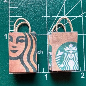 May include: Two miniature paper shopping bags with rope handles. One bag features the Starbucks logo with a green and brown color scheme. The other bag has the Starbucks logo with a green and brown color scheme. The bags are on a green cutting mat.