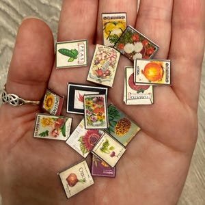 May include: A collection of miniature rectangular seed packets featuring various colorful illustrations of vegetables and flowers, including peas, tomatoes, onions, and sunflowers, with printed text labels on each individual small paper packet.