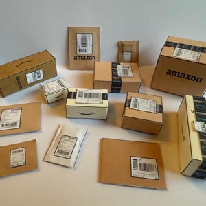 May include: An assortment of Amazon-branded cardboard boxes and packaging, including boxes of various sizes, envelopes, and a small padded mailer. The boxes have black tape and the Amazon logo. Some boxes have the word "prime" printed on them.