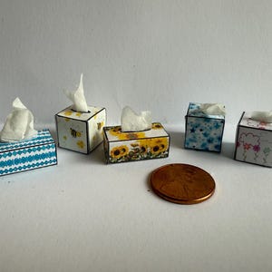May include: Miniature tissue boxes with various designs, including blue geometric patterns, sunflowers, bees, and floral prints. Each box has a white tissue sticking out. A penny is next to the boxes for scale.