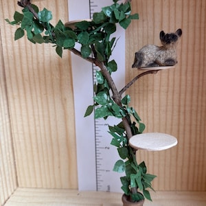 May include: A miniature cat tree with three platforms and a small, brown cat figurine sitting on one of the platforms. The tree is made of brown branches and has green leaves. The tree is sitting on a white square base.