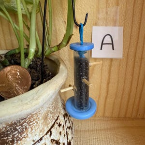 May include: A small, blue and clear bird feeder filled with black seeds hangs from a black metal hook. The feeder is attached to a blue base and top. A potted plant and a penny are in the background. The letter "A" is on a white square.