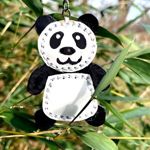 May include: A black and white panda bear keychain made of leather with stitched details. The panda is hanging from a silver chain against a green leafy background.