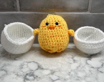 May include: A yellow crocheted chick toy with black eyes and an orange beak, standing between two white crocheted egg-shaped baskets.