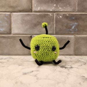 May include: A green crocheted plush toy with black pipe cleaner arms and legs. The toy has black eyes and pink cheeks.