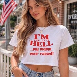 May include: White crop top with the text "I'M THE ONLY HELL...my mama ever raised!" in red. The shirt is paired with blue jeans. An American flag is visible in the background.