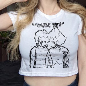 May include: White crop top with a black graphic of two figures and the text "PG.99 and CITY OF CATERPILLAR A SPLIT PERSONALITY". The shirt is made of a soft material and has short sleeves.