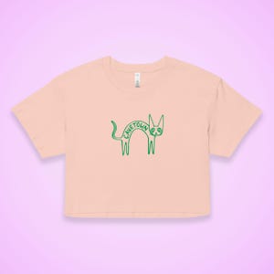 May include: A light peach-colored crop top with a green outline of a cat. The word "CAVETOWN" is written in green across the cat's body. The shirt has short sleeves and a crew neck.