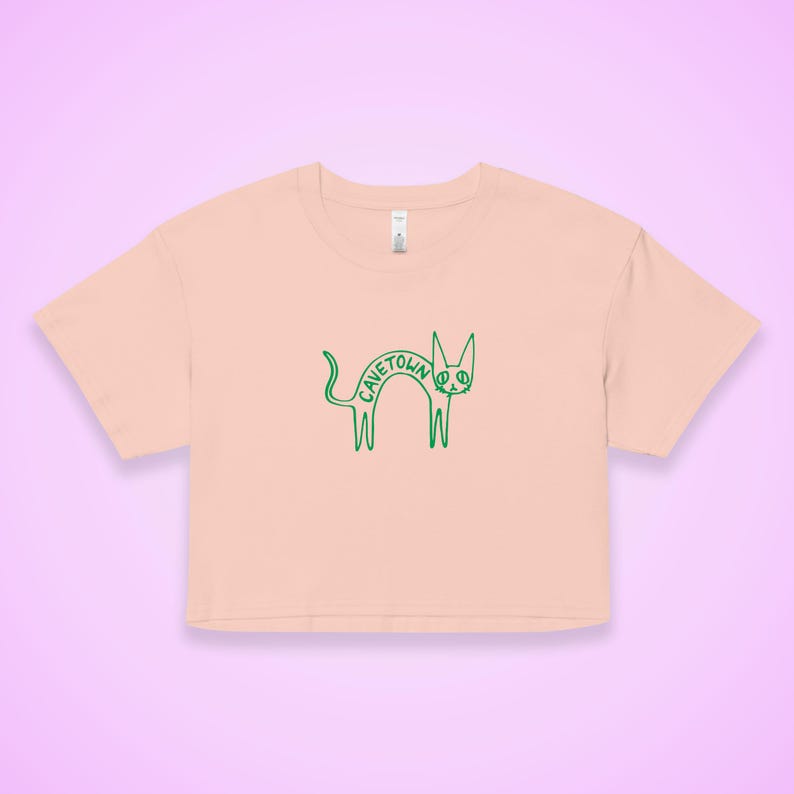 May include: A light peach-colored crop top with a green outline of a cat. The word "CAVETOWN" is written in green across the cat's body. The shirt has short sleeves and a crew neck.