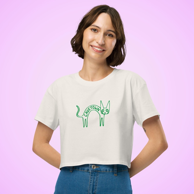 May include: Cream-colored crop top with a green cat graphic and the word "CAVETOWN" arched over the cat's back. The cat design is a simple, whimsical line drawing. The shirt has short sleeves and a crew neck.