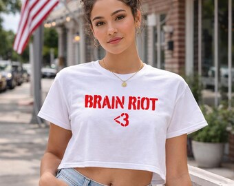 May include: White crop top with the red text "BRAIN RIOT" and a heart symbol. The shirt is paired with blue jeans. An American flag is visible in the background.