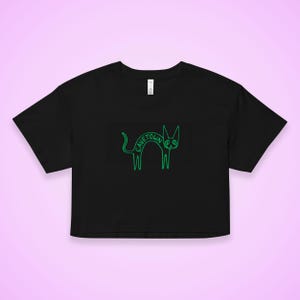 May include: Black crop top with a neon green cat graphic. The cat illustration has the word "CAVETOWN" written across its body. The shirt has a crew neck and short sleeves. The background is a light purple.