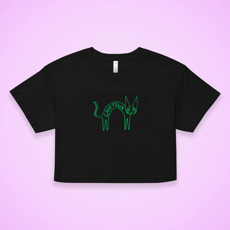 May include: Black crop top with a neon green cat graphic. The cat illustration has the word "CAVETOWN" written across its body. The shirt has a crew neck and short sleeves. The background is a light purple.