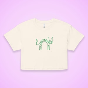May include: Cream-colored crop top with a green outline drawing of a cat. The cat's body spells out the word "CAVETOWN". The shirt has short sleeves and a crew neck.