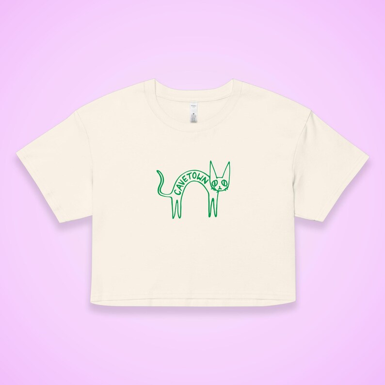 May include: Cream-colored crop top with a green outline drawing of a cat. The cat's body spells out the word "CAVETOWN". The shirt has short sleeves and a crew neck.