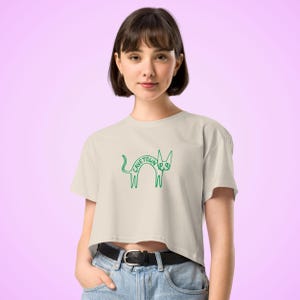 May include: Cream-colored crop top with a green cat graphic and the text "CAVETOWN". The graphic features a stylized cat with the band's name. The shirt has short sleeves and a crew neck.