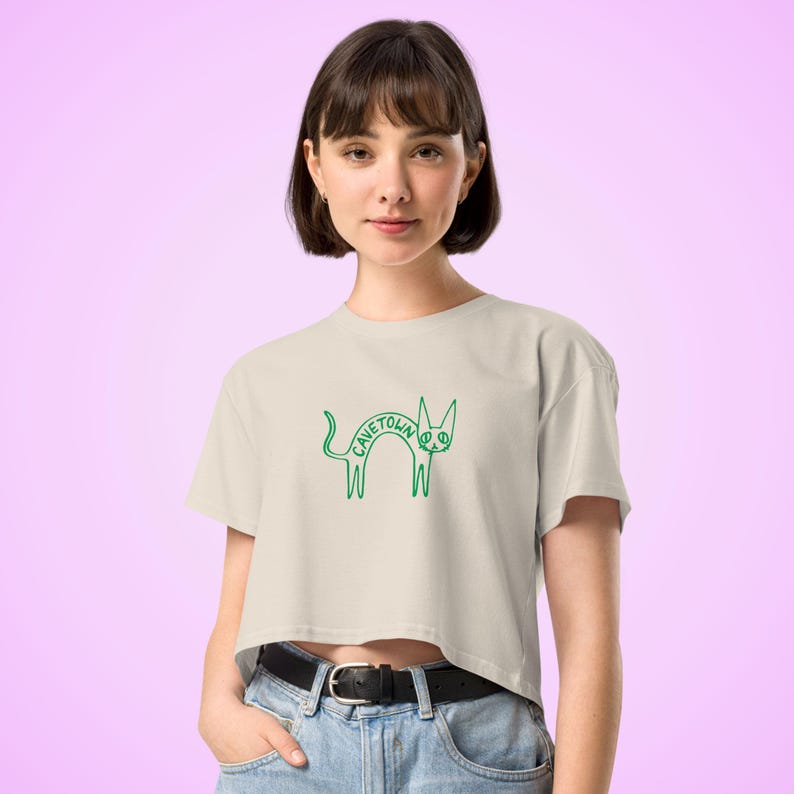 May include: Cream-colored crop top with a green cat graphic and the text "CAVETOWN". The graphic features a stylized cat with the band's name. The shirt has short sleeves and a crew neck.