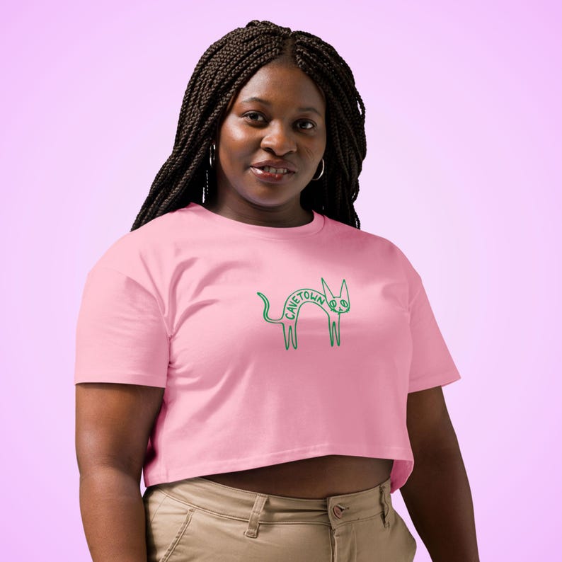 May include: A light pink crop top with a green outline of a cat and the word "CAVETOWN" in green. The shirt is made of a soft material and has short sleeves.
