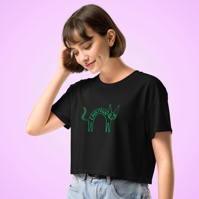May include: Black crop top with a green outline drawing of a cat with the word "CAVETOWN" arched across its body. The cat has a peace sign for an eye. The shirt has short sleeves and a crew neck.